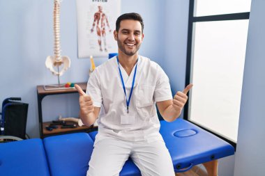 Sakallı, sakallı, acılı klinikte çalışan genç bir adam. Elleriyle olumlu jest yapıyor. Gülümsüyor ve mutlu. Neşeli ifade ve kazanan jest. 