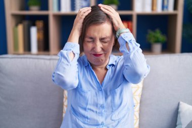 Orta yaşlı İspanyol bir kadın evdeki kanepede oturup acı ve migren yüzünden çaresiz ve stresli bir şekilde baş ağrısı çekiyor. Eller başının üstünde.. 