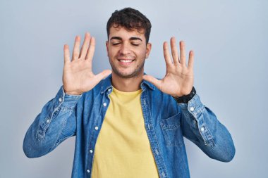 Young hispanic man standing over blue background showing and pointing up with fingers number ten while smiling confident and happy. 