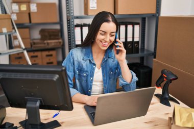 Young beautiful hispanic woman ecommerce business worker using laptop talking on smartphone at office