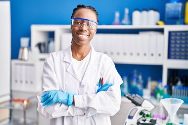 Afrikalı Amerikalı bilim kadını gülümsüyor. Kollarını kavuşturup laboratuvarda duruyor.