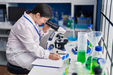 Down sendromlu genç bir kadın. Laboratuvarda mikroskop kullanarak belgelere yazı yazıyor.