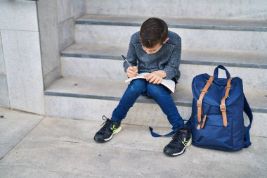 Blond child student writing on book sitting on stairs at school