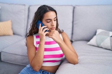 Young beautiful hispanic woman talking on smartphone with worried expression at home