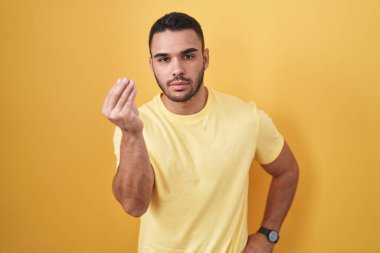 Young hispanic man standing over yellow background doing italian gesture with hand and fingers confident expression 