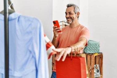 Orta yaşlı, gri saçlı, güler yüzlü, kendine güvenen bir müşteri elbise mağazasındaki akıllı telefondan kıyafetlere fotoğraf çeker.