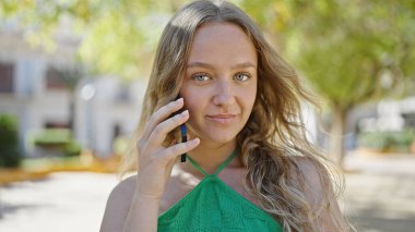 Young blonde woman talking on smartphone with serious expression at park