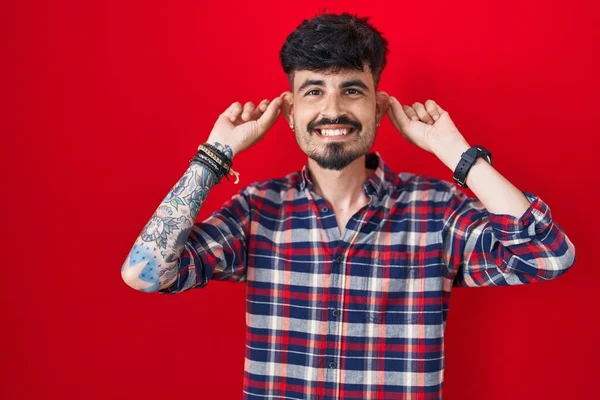 髭を生やした若いヒスパニック系の男は 赤い背景に指で耳を引く笑みを浮かべて立って 面白いジェスチャー オーディション問題 — ストック写真
