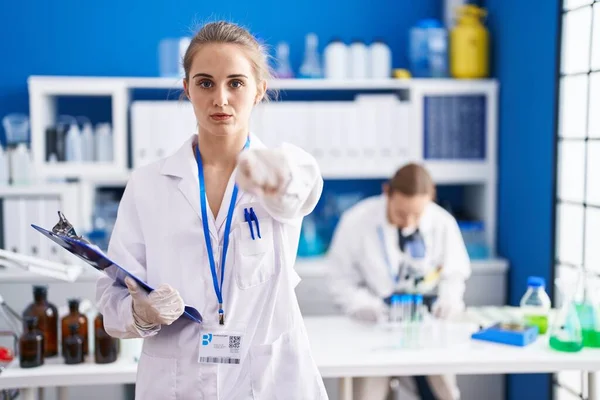 Blonde woman working at scientist laboratory pointing with finger to the camera and to you, confident gesture looking serious