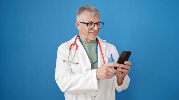 中年白髪の男医師笑顔自信を持って使用してスマートフォン上の隔離された青の背景 — ストック写真