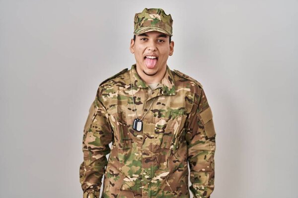 Young arab man wearing camouflage army uniform sticking tongue out happy with funny expression. emotion concept. 