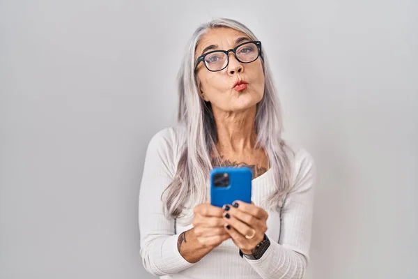 中年妇女灰白的头发使用智能手机打字信息看着相机吹一个吻是可爱和性感的 爱的表达 — 图库照片
