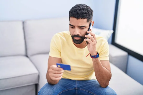 stock image Young arab man talking on smartphone and credit card sitting on sofa at home