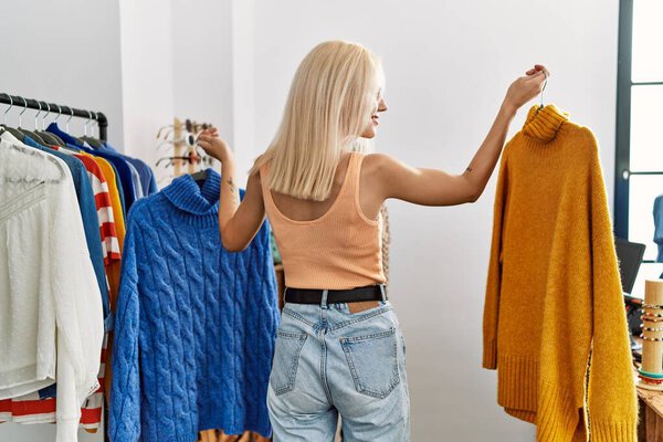 Young blonde woman customer choosing sweater standing backwards at clothing store