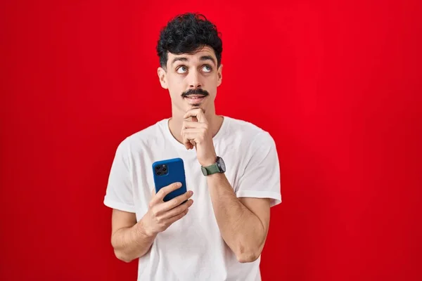Homem Hispânico Usando Smartphone Sobre Fundo Vermelho Com Mão Queixo — Fotografia de Stock