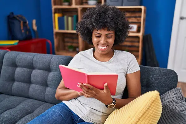 Evdeki kanepede oturan Afro-Amerikalı kadın kitap okuyor.