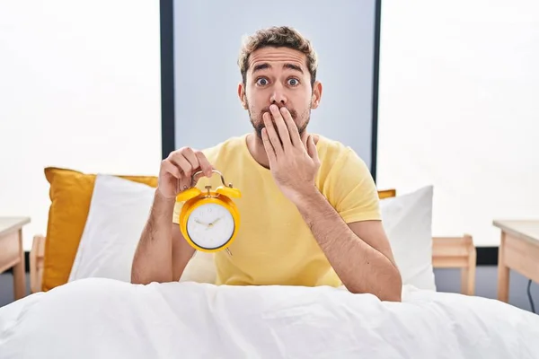 Hispanic Man Beard Holding Alarm Clock Bed Covering Mouth Hand — Stock fotografie