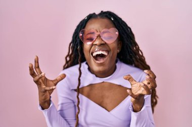 Pembe arka planda örülü saçları olan Afrikalı bir kadın. Deli gibi bağırıyor, agresif bir ifadeyle bağırıyor ve kollarını kaldırıyor. hüsran kavramı. 