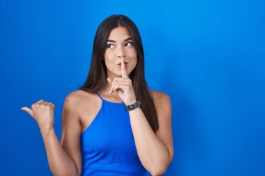 Mavi arka planda dikilen İspanyol kadın sessiz olmak istiyor. Parmakları dudaklarında, elleri yandan işaret ederek. sessizlik ve gizli konsept. 