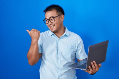 Çinli genç adam bilgisayarını kullanarak gülümsüyor, gülümsüyor ve başparmağıyla yan tarafa bakıyor.. 