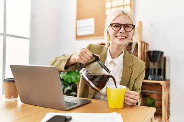 Genç sarışın iş kadını laptopuyla ofisteki bardağa kahve dolduruyor.