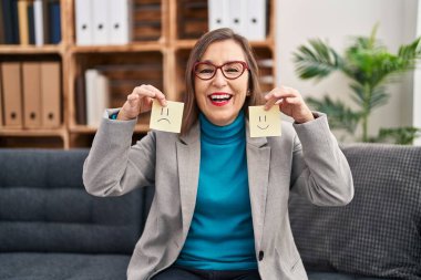Middle age hispanic woman working on depression holding sad to happy emotion paper smiling and laughing hard out loud because funny crazy joke. 