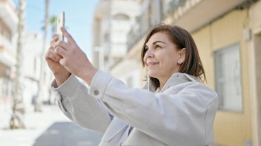 Orta yaşlı, kendine güvenen İspanyol bir kadın sokakta akıllı telefonuyla fotoğraf çekiyor.