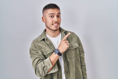 Genç İspanyol adam izole edilmiş arka planda neşeli bir şekilde ayakta duruyor. Yüzünde mutlu ve doğal bir ifadeyle elleri ve parmaklarıyla işaret eden bir gülümseme var. 