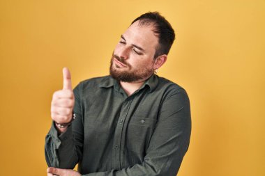 Plus size hispanic man with beard standing over yellow background looking proud, smiling doing thumbs up gesture to the side 