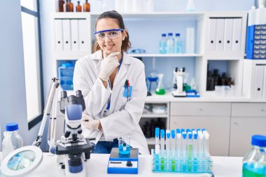 Young hispanic woman working at scientist laboratory looking confident at the camera smiling with crossed arms and hand raised on chin. thinking positive. 