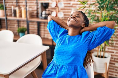 Afro-Amerikalı kadın ellerini başının üstüne koyup evinde sandalyede oturuyor.