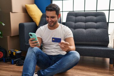 Young hispanic man using smartphone and credit card sitting on floor at new home