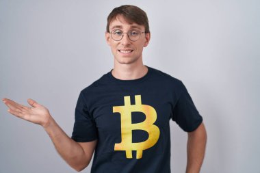 Beyaz, sarışın, bitcoin tişörtlü, neşeli gülümseyen, elinde avucuyla kameraya bakan bir adam.. 