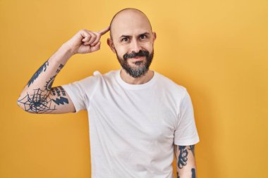 Sarı arka planda dikilen dövmeleri olan genç İspanyol bir adam tek parmağıyla kafasını gösteren, harika bir fikir ya da düşünce, iyi bir hafıza. 