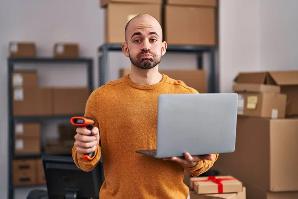 Young bald man with beard working at small business ecommerce with laptop puffing cheeks with funny face. mouth inflated with air, catching air.