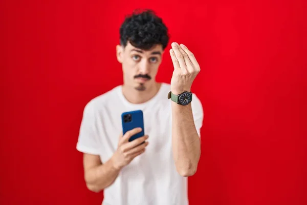 Homem Hispânico Usando Smartphone Sobre Fundo Vermelho Fazendo Gesto Italiano — Fotografia de Stock