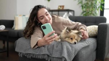 Köpekli genç İspanyol kadın evdeki kanepede uzanmış video görüşmesi yapıyor.