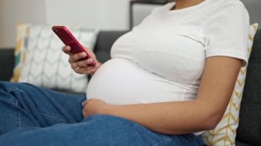 Evdeki kanepede oturan akıllı telefon kullanan hamile bir kadın.