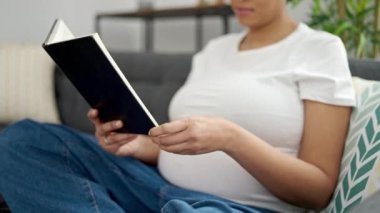 Young pregnant woman reading book sitting on sofa at home
