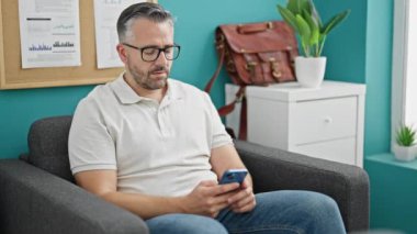 Gri saçlı iş adamı akıllı telefon kullanarak ofise bakıyor.