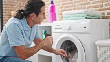 Çamaşırhanede çamaşır yıkayan orta yaşlı bir adam akıllı telefon tutuyor.