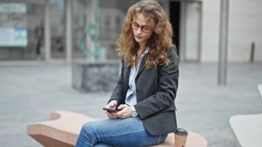 Akıllı telefon kullanan genç bir iş kadını caddede bankta oturuyor.