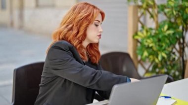 Genç, kızıl saçlı, iş kadını. Laptop kullanıyor. Kahve dükkanının terasında not alıyor.