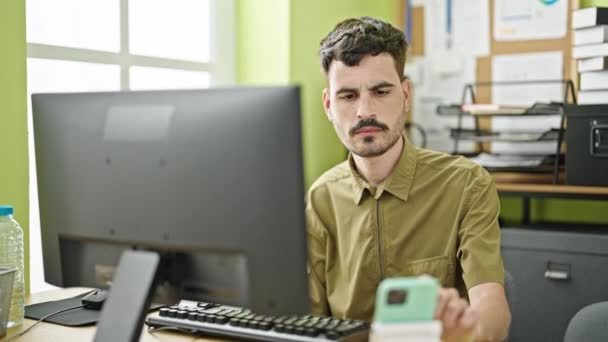 オフィスで働くコンピュータおよびスマートフォンを使用して若いヒスパニックの男のビジネスワーカー — ストック動画