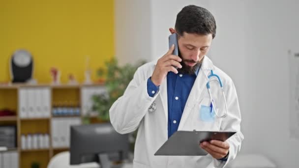Young Hispanic Man Doctor Reading Document Clipboard Talking Smartphone Clinic — Stock Video