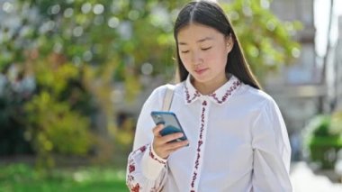 Parkta akıllı telefondan müzik dinleyen Çinli genç bir kadın.