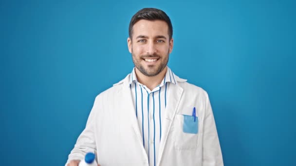 若いヒスパニックの男性医師は 孤立した青い背景の上に薬瓶を保持する自信を持って微笑みます — ストック動画