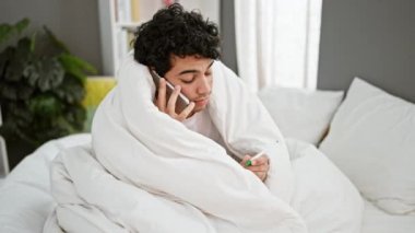 Battaniye tutan genç latin adam yatak odasında akıllı telefondan konuşuyor.