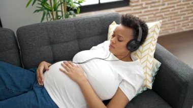 Genç hamile kadın evdeki kanepede müzik dinliyor.