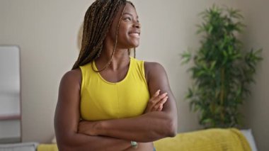 Afro-Amerikalı kadın gülümsüyor. Kollarını kavuşturup evde duruyor.